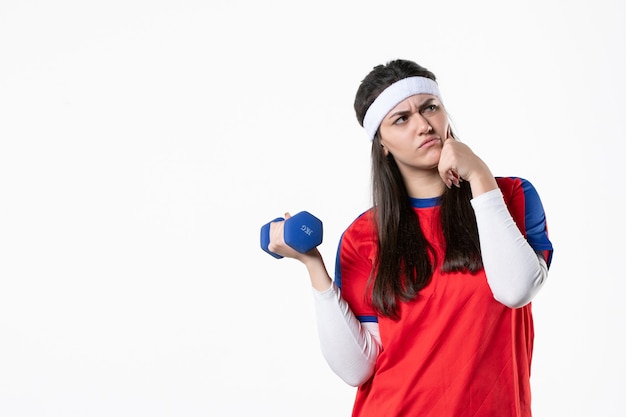 Vue de face jeune femme en vêtements de sport avec des haltères bleus sur un mur blanc