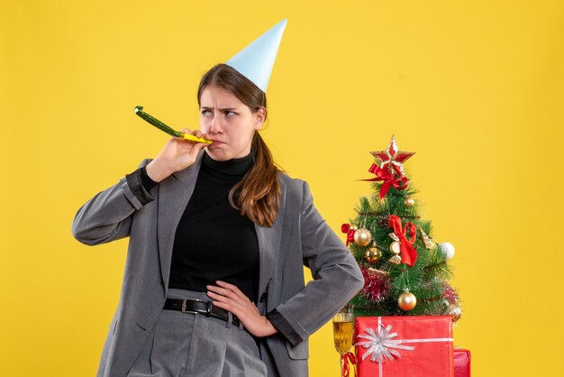 Vue de face jeune fille avec chapeau de fête à l'aide de bruiteur debout près de l'arbre de Noël et des cadeaux cocktail