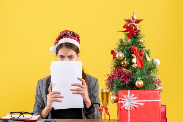 Vue de face jeune fille avec chapeau de Noël assis à la table couvrant son visage avec des documents arbre de Noël et des cadeaux cocktail