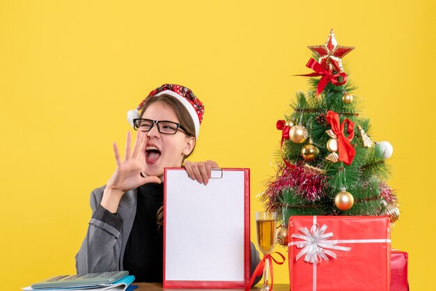 Vue de face jeune fille avec chapeau de Noël assis à la table en criant arbre de Noël et cadeaux cocktail