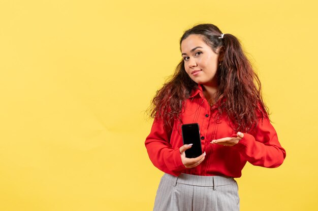 Vue de face jeune fille en chemisier rouge avec des cheveux mignons tenant son téléphone sur fond jaune couleur enfant enfant fille jeunesse innocence