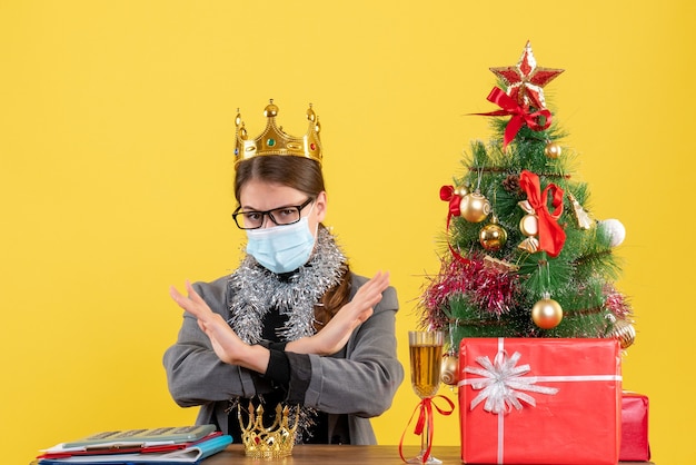 Vue de face jeune fille avec couronne portant masque et lunettes croisant ses mains arbre de Noël et cadeaux cocktail