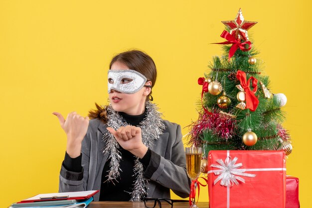 Vue de face jeune fille avec masque assis à la table montrant quelque chose avec un arbre de Noël doigt et un cocktail de cadeaux