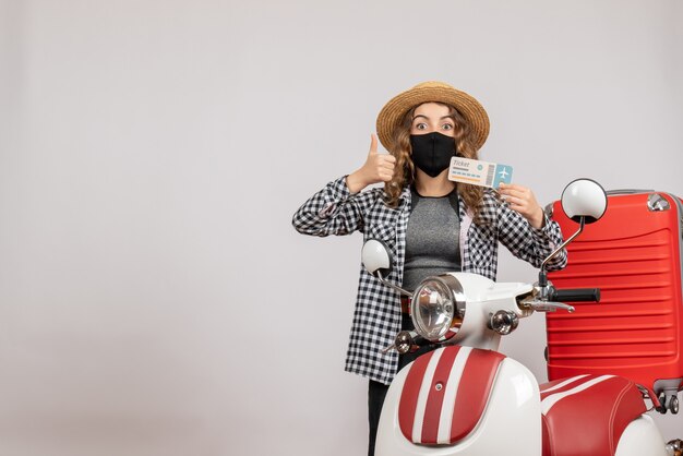 Vue de face jeune fille avec un masque noir tenant un billet donnant le pouce levé debout près d'un cyclomoteur rouge