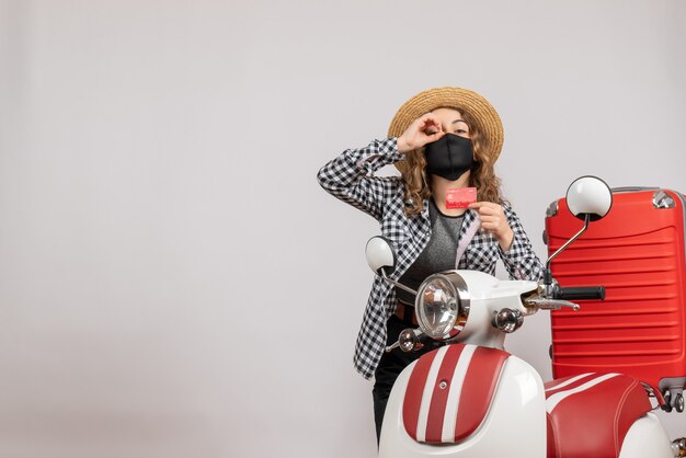 Vue de face jeune fille avec masque noir tenant un billet faisant des jumelles à main debout près d'un cyclomoteur rouge