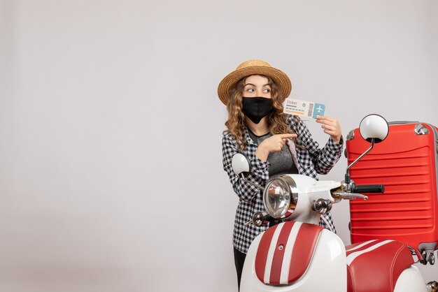 Vue de face jeune fille avec masque noir tenant un billet pointant vers la droite près d'un cyclomoteur rouge