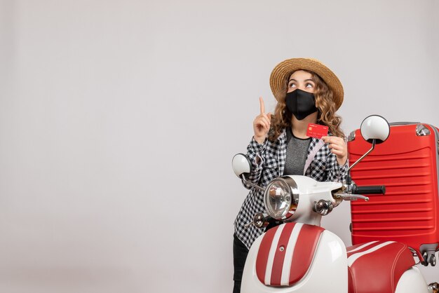 Vue de face jeune fille avec masque noir tenant un billet pointant vers le haut, debout près d'un cyclomoteur rouge