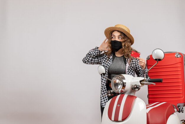 Vue de face jeune fille avec masque noir tenant une carte écoutant quelque chose debout près d'un cyclomoteur rouge