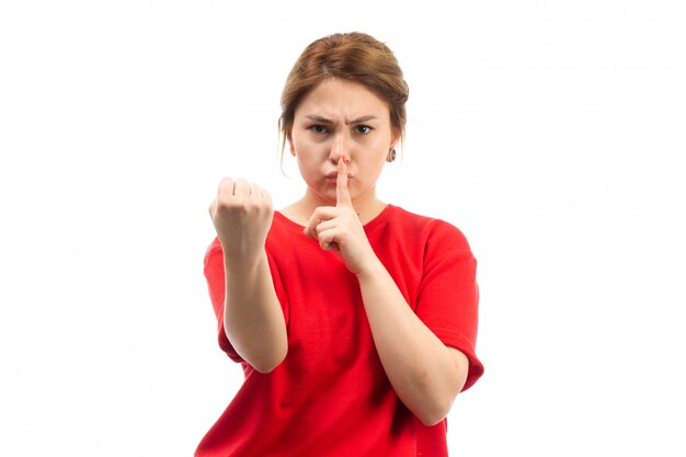 Une vue de face jeune fille séduisante en t-shirt rouge portant un jean noir menaçant le silence avec le poing sur le blanc