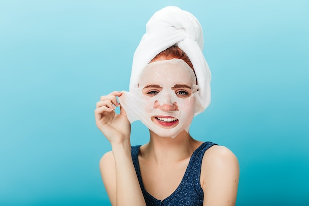 Vue de face d'une jeune fille souriante décollant le masque facial. Photo de Studio de femme heureuse avec une serviette sur la tête posant sur fond bleu.