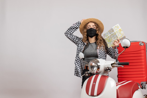 Vue de face jeune fille voyageuse avec masque noir tenant une carte debout près d'un cyclomoteur rouge