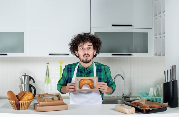 Vue de face d'un jeune homme ambitieux tenant une pâtisserie fraîchement préparée dans une petite boîte dans la cuisine blanche