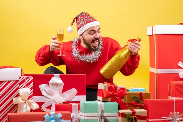 Vue de face jeune homme assis autour de cadeaux avec du champagne sur fond jaune