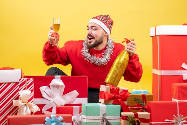 Vue de face jeune homme assis autour de cadeaux avec du champagne sur fond jaune