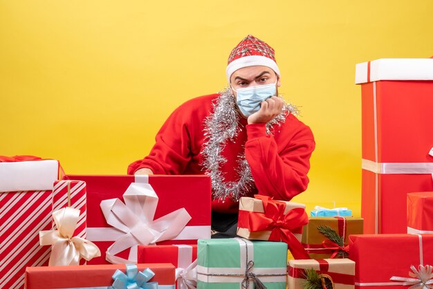 Vue de face jeune homme assis autour de cadeaux en masque sur fond jaune