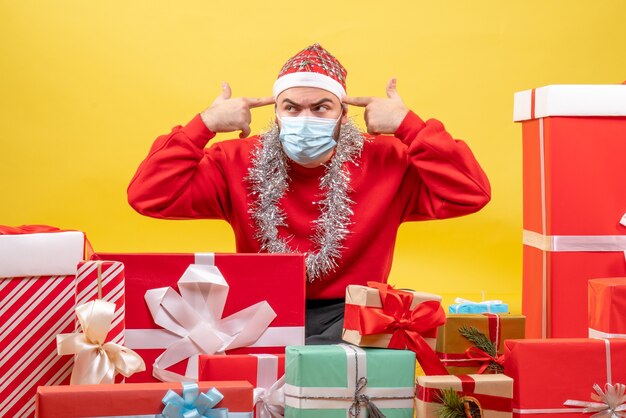 Vue de face jeune homme assis autour de cadeaux en masque sur fond jaune