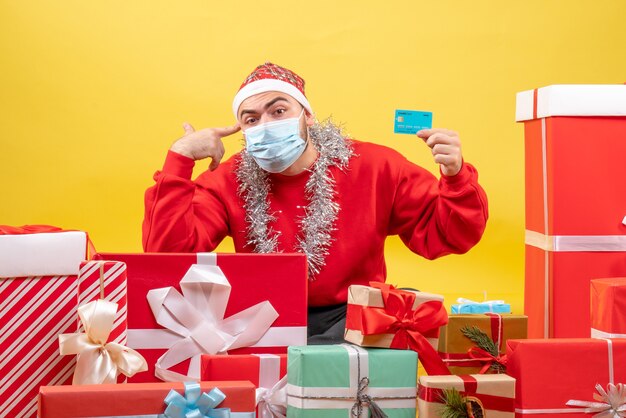 Vue de face jeune homme assis autour de cadeaux en masque stérile sur fond jaune