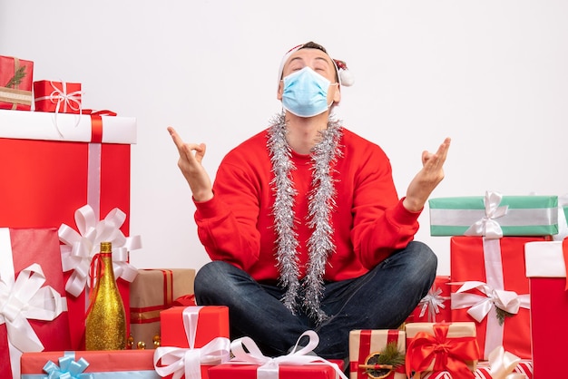 Vue de face d'un jeune homme assis autour de cadeaux méditant sur un mur blanc