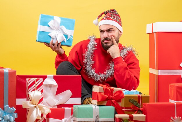 Vue de face jeune homme assis autour de cadeaux de Noël sur fond jaune