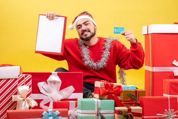 Vue de face jeune homme assis autour de cadeaux de Noël sur fond jaune