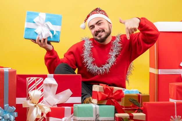 Vue de face jeune homme assis autour de cadeaux de Noël sur fond jaune