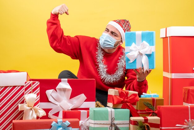 Vue de face jeune homme assis autour de cadeaux de Noël sur jaune