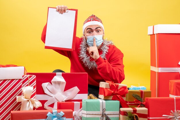 Vue de face jeune homme assis autour de cadeaux de Noël avec note sur fond jaune