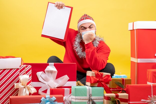 Vue de face jeune homme assis autour de cadeaux de Noël avec note sur jaune
