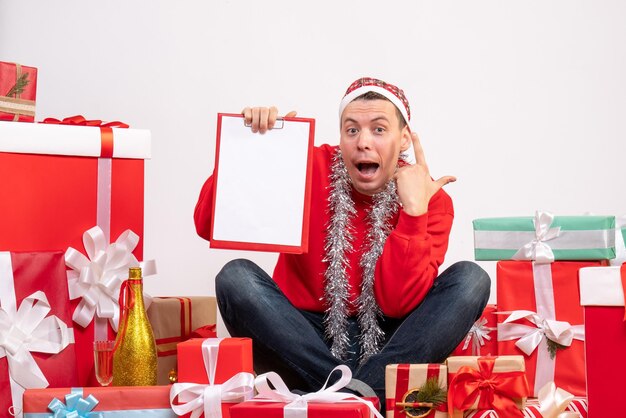 Vue de face d'un jeune homme assis autour de cadeaux de Noël avec une note sur le mur blanc