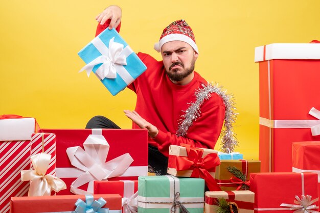 Vue de face jeune homme assis autour de cadeaux et en tenant un sur fond jaune