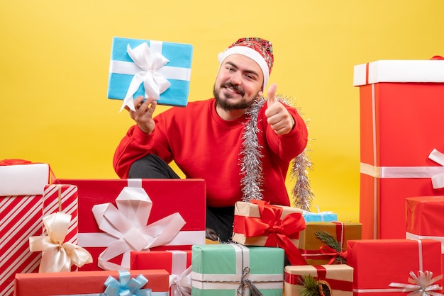 Vue de face jeune homme assis autour de cadeaux et en tenant un sur fond jaune