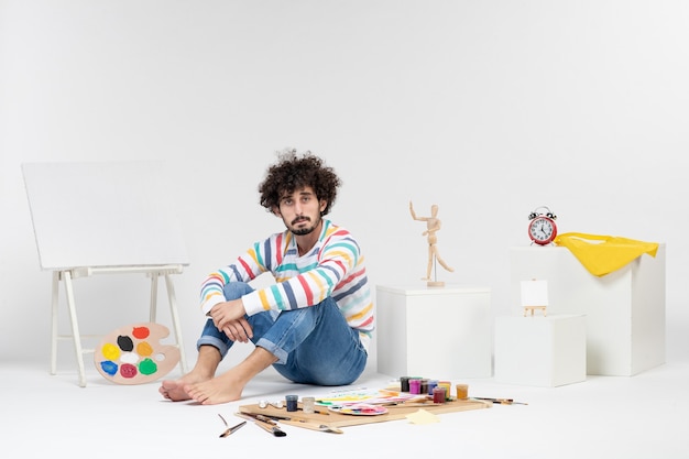 Vue de face d'un jeune homme assis autour de peintures et de dessins ennuyés sur un mur blanc