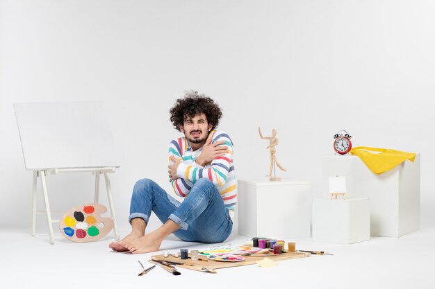 Photo gratuite vue de face d'un jeune homme assis autour de peintures et de dessins sur un mur blanc