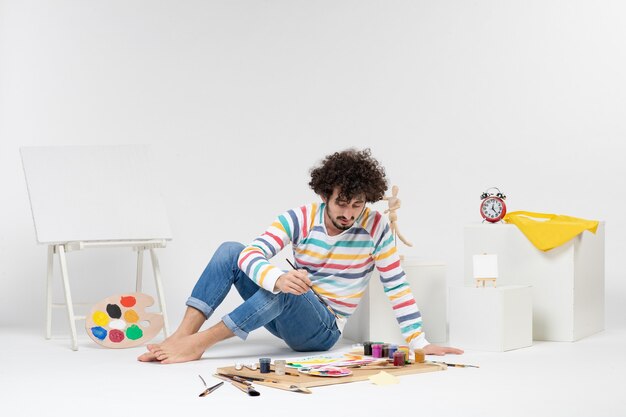 Vue de face d'un jeune homme assis autour de peintures et de dessins sur un mur blanc