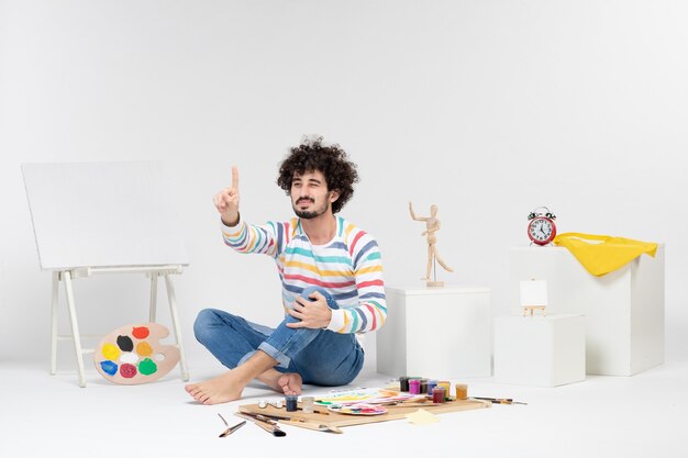 Vue de face d'un jeune homme assis autour de peintures et de dessins sur un mur blanc