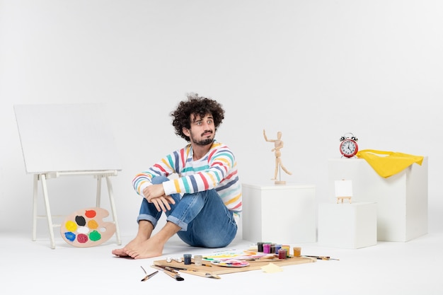 Vue de face d'un jeune homme assis autour de peintures et de dessins sur un mur blanc