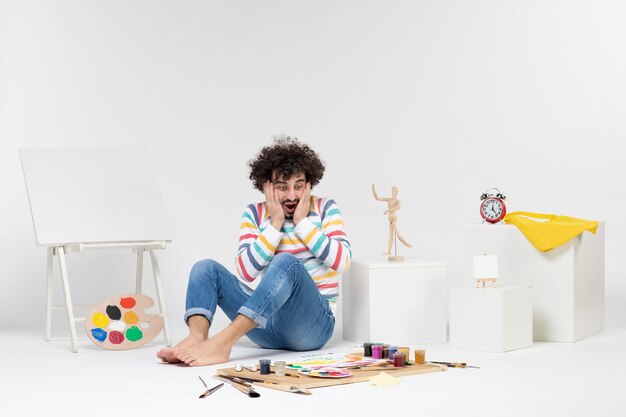 Vue de face d'un jeune homme assis autour de peintures et de dessins sur un mur blanc