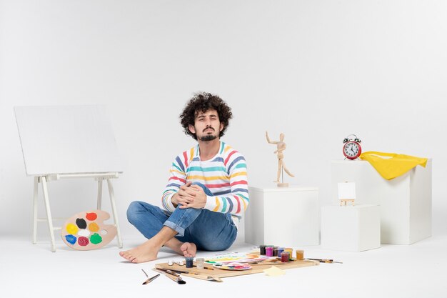 Vue de face d'un jeune homme assis autour de peintures et de dessins sur un mur blanc