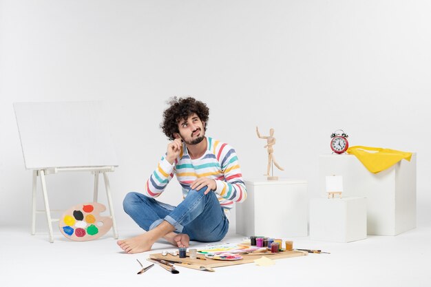 Vue de face d'un jeune homme assis autour de peintures et de dessins sur un mur blanc