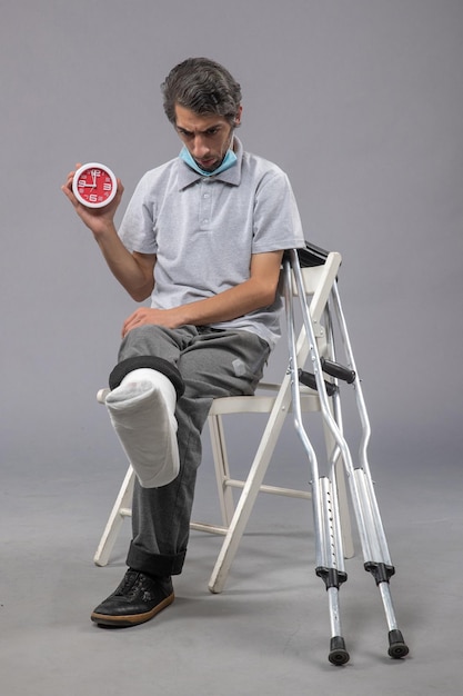 Photo gratuite vue de face jeune homme assis avec un bandage attaché au pied cassé et tenant une horloge sur un bureau gris torsion douleur pied accident jambe mâle