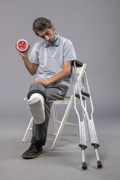 Vue de face jeune homme assis avec un bandage attaché au pied cassé et tenant une horloge sur un mur gris douleur torsion du pied accident jambe mâle