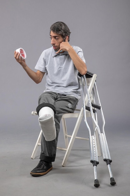 Photo gratuite vue de face jeune homme assis avec un bandage attaché au pied cassé et tenant une horloge sur le mur gris pied torsion mâle accident douleur jambe