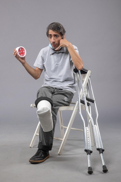 Vue de face jeune homme assis avec un bandage attaché au pied cassé et tenant une horloge sur le mur gris pied torsion mâle accident douleur jambe