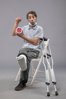 Vue de face jeune homme assis avec un bandage attaché en raison d'un pied cassé et tenant une horloge sur le mur gris torsion de la jambe douleur au pied accident mâle