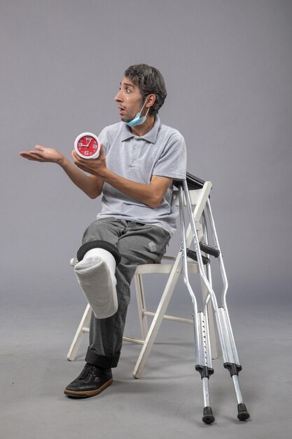 Vue de face jeune homme assis avec un bandage attaché en raison d'un pied cassé et tenant une horloge sur un mur gris torsion des jambes douleur au pied accident mâle