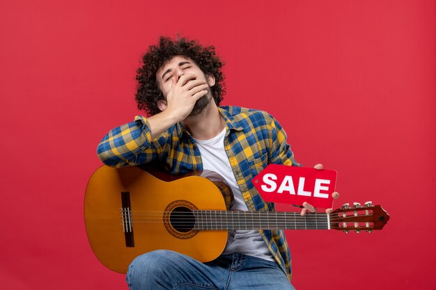 Vue de face jeune homme assis avec guitare bâillant sur le mur rouge concert live applaudissements musicien vente jouer couleur