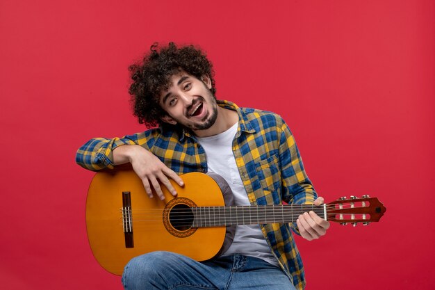 Vue de face jeune homme assis avec guitare sur mur rouge concert live bande de couleur jouer des applaudissements des musiciens