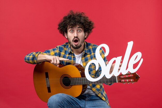 Vue de face jeune homme assis avec guitare sur mur rouge concert live musicien vente musique couleur applaudissements