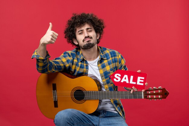 Vue de face jeune homme assis avec guitare sur mur rouge concert live musique couleur applaudissements musicien vente