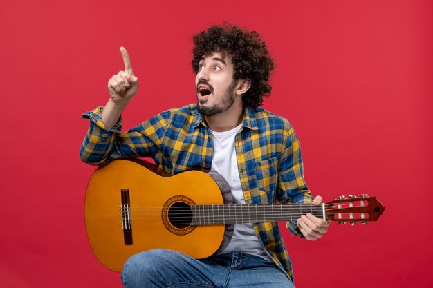 Vue de face jeune homme assis avec guitare sur mur rouge jouer concert live band musique couleurs musicien applaudissements